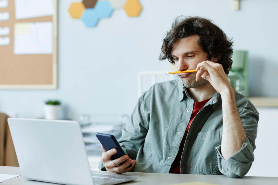 psychology  Portrait of Caucasian young man playing with pencil and using phone at workplace suffering from boredom and procrastination