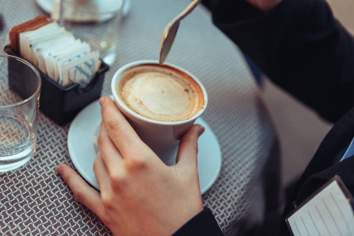 A person stirs a cup of coffee with a spoon while sitting at a table with a glass of water and a container holding sugar packets