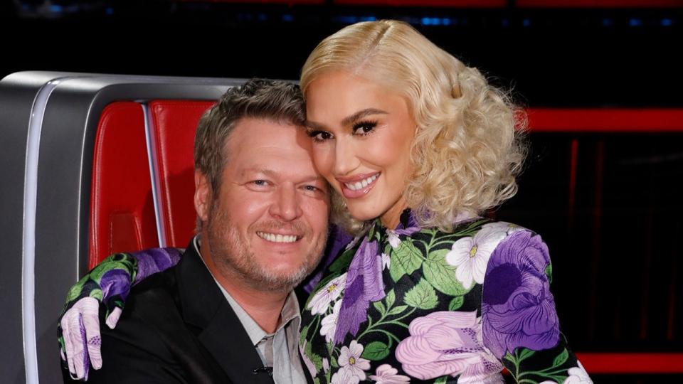 Blake Shelton, wearing a black jacket and shirt, hugs wife Gwen Stefani in a purple, green and white floral outfit.