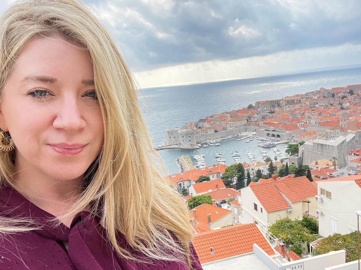 A blonde woman taking a selfie in front of red roof buildings in Dubrovnik, Croatia.
