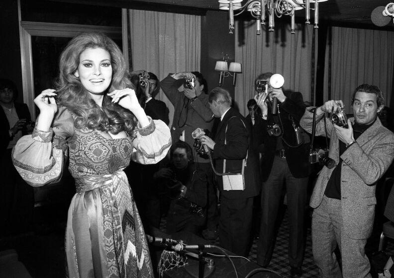 A black-and-white photo of a woman in a dress with journalists to her right taking photos