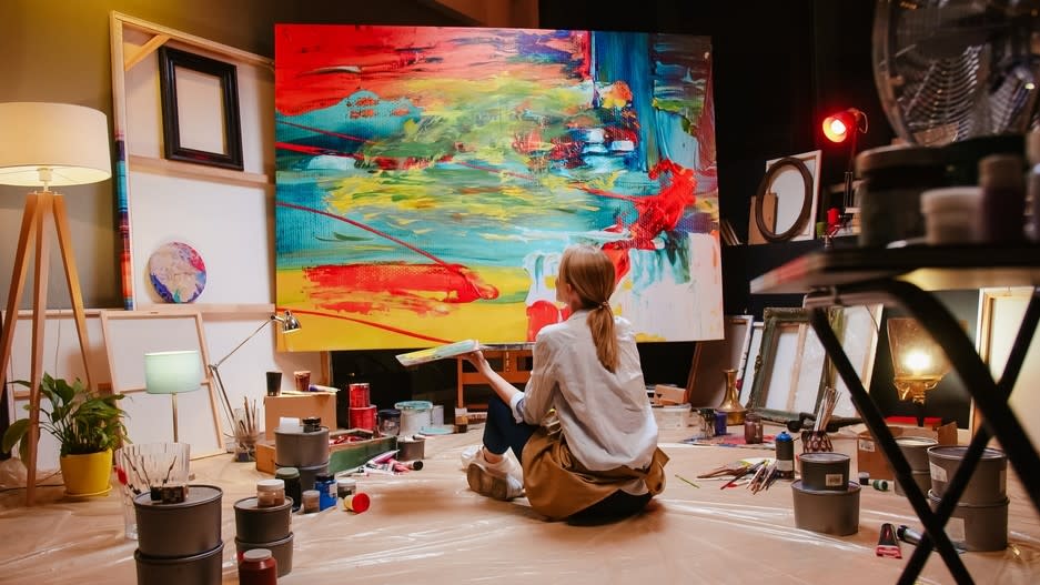 A woman sits on the floor in a studio in front of a painting.