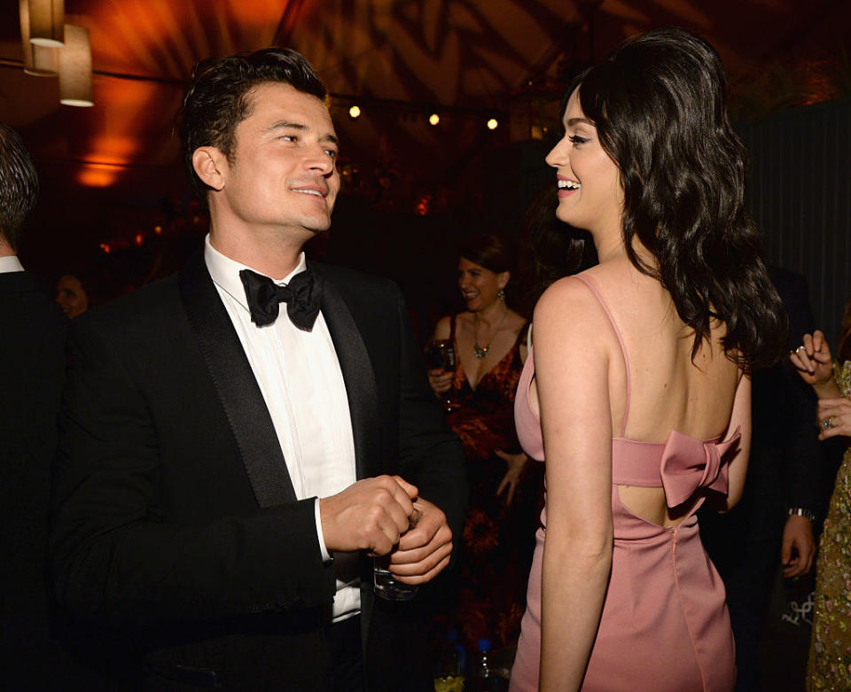 Orlando Bloom in a tuxedo and Katy Perry in a dress with bow accents conversing at an event