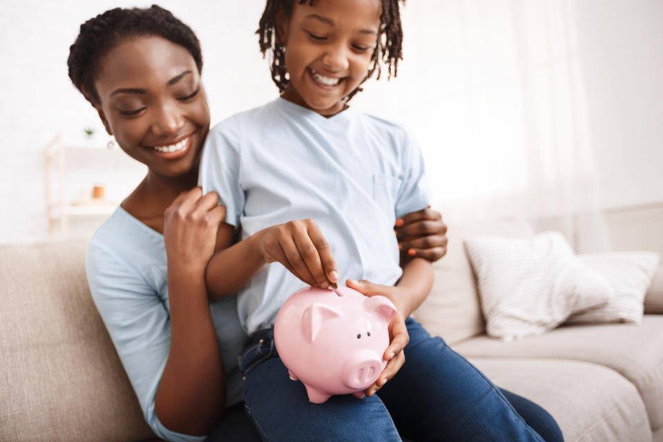 Home Finances Concept. Happy african kid putting coins into piggy bank, sitting on mom&#39;s lap, copyspace