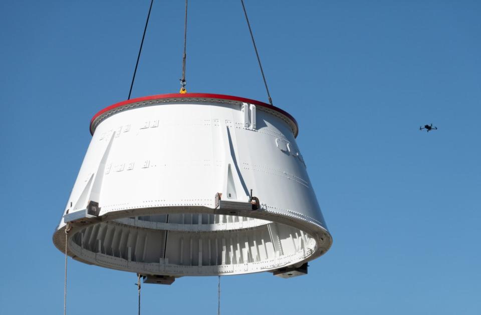 A solid socket booster aft skirt is lifted in the air with sky and a small drone flying at left as backdrop