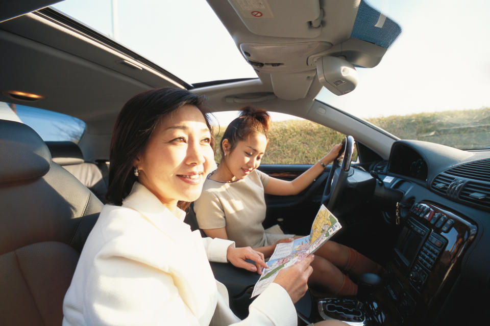 Two women in a car, one at the wheel and the other reading a map, appear ready for a road trip