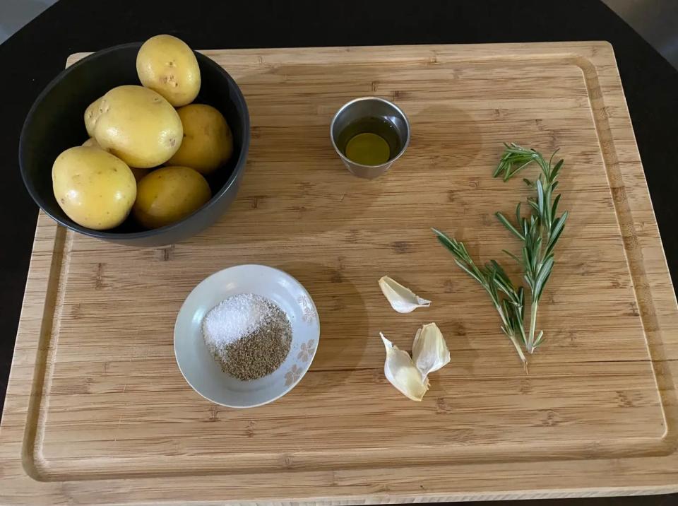 potatoes, oil, seasoning, garlic cloves, and rosemary on a wooden cutting board