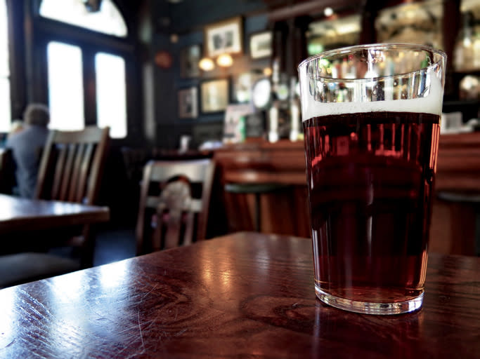 A clear pint of beer sits on a wooden table in a cozy pub setting with chairs and framed pictures visible in the background