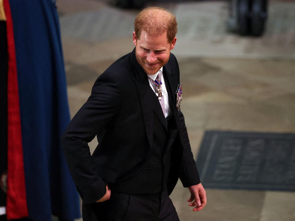 Prince Harry walking in the interior of Westminster Abbey with one hand in the pocket of his black suit.