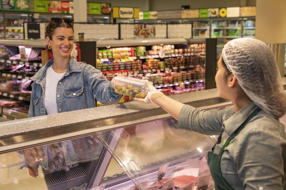 Woolworths worker hands a customer a plastic container of olives.