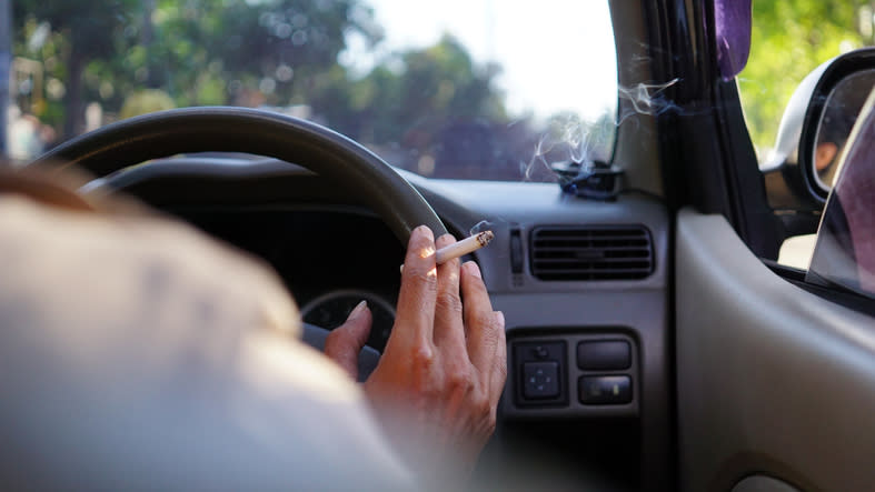 Pictured is a driver holding the steering wheel with a cigarette between their fingers. 