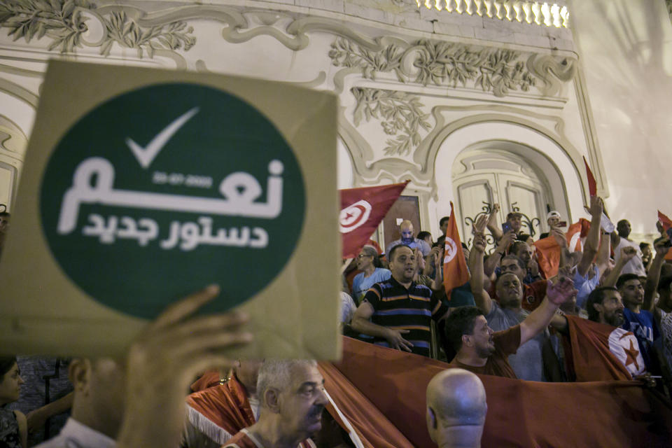 FILE - A Tunisian holds a sign reading 'Yes to the new constitution' as others celebrate exit polls that indicate a vote in favor of the new constitution, in Tunis, July 25, 2022. Tunisians are expressing a range of reactions to the adoption of a new constitution. Voters overwhelmingly supported the document in a referendum on Monday, July 25 although the vote was marked by low turnout. The new political system will see sweeping executive powers given to the president and the removal of key checks and balances, causing critics to warn that Tunisia may see a reversal of hard-won democratic gains. (AP Photo/Riadh Dridi, file)