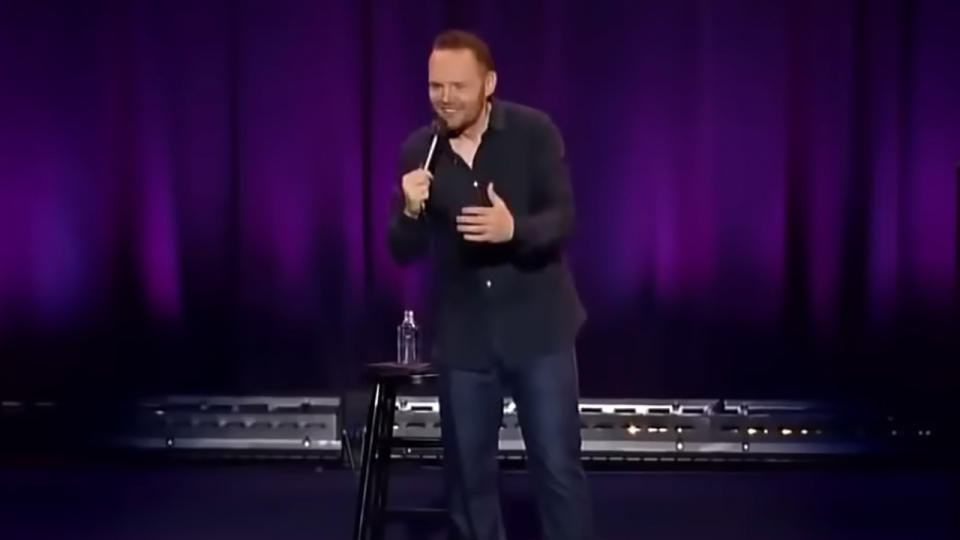 Bill Burr smiling, wearing black, on stage with a microphone