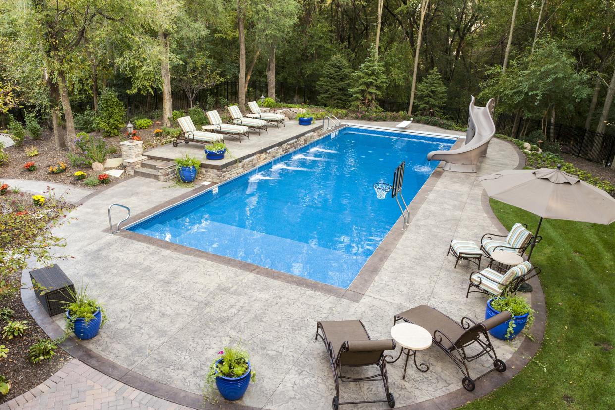 Large in-ground swimming pool with a slide, a basketball hoop, and a patio in a secluded backyard surrounded by trees