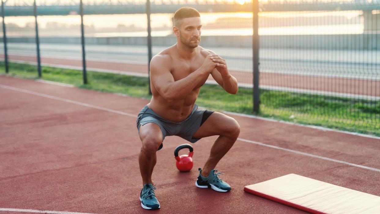  Man performing a squat outdoors next to exercise mat and kettlebell. 