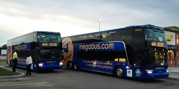 Two side-by-side Megabuses during a pitstop. Two people are standing besides the first bus parked further back.