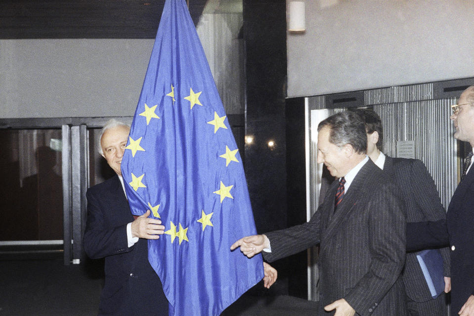 FILE - Soviet foreign minister Eduard Shevardnadze peeking from behind the European Community flag, while EC Commission President Jacques Delors points to the spot where he should be standing for the picture session, at the Commission's head office in Brussels, Belgium, on Dec. 18, 1989. Delors, a Paris bank messenger’s son who became the visionary and builder of a more unified Europe in his momentous decade as chief executive of the European Union, has died in Paris, his daughter Martine Aubry said Wednesday Dec. 27, 2023. He was 98. (AP Photo/Carl Duyck, File)