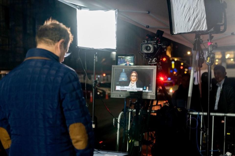 a man with his back to the camera looks onward at a tv screen with a news broadcaster and camera lights