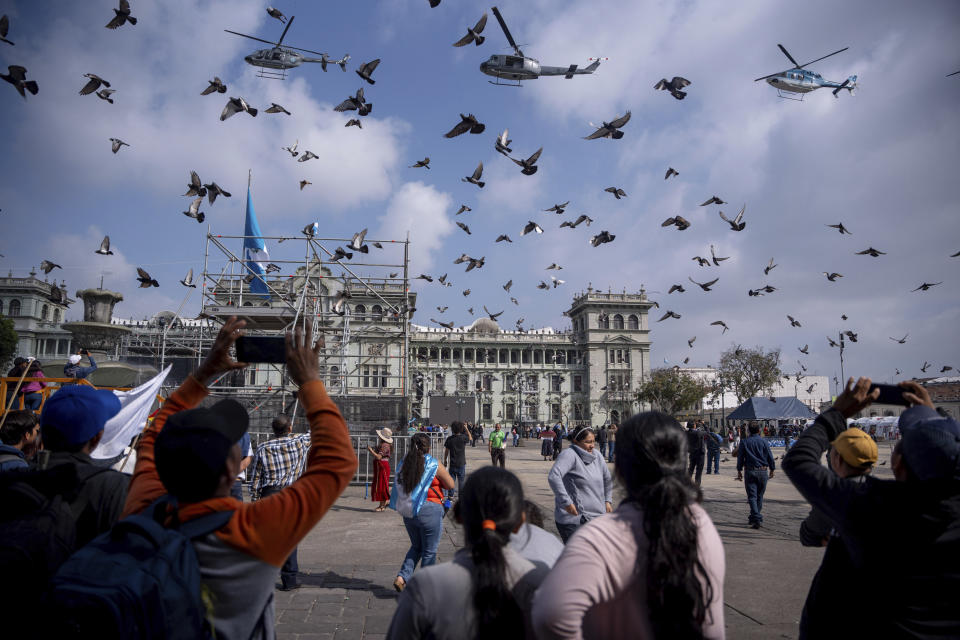 Varias personas mirando helicópteros en formación sobre la plaza de la Constitución antes de la investidura del presidente electo de Guatemala Bernardo Arévalo, en el entorno del Palacio Nacional en Ciudad de Guatemala, el domingo 14 de enero de 2024. (AP Foto/Santiago Billy)