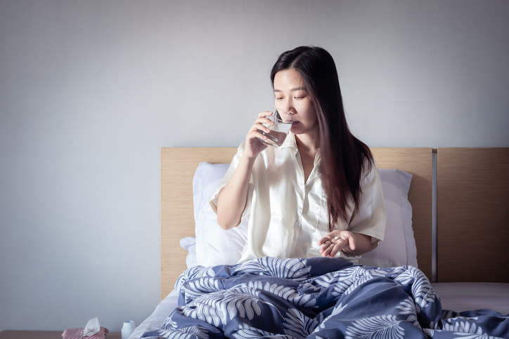 A person sitting in bed and taking a sip of water to go with their pills