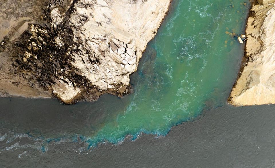 Green water, seen from above, flowing from hard, dry land into dark, muddy water, turning blue at the edges where they mingle