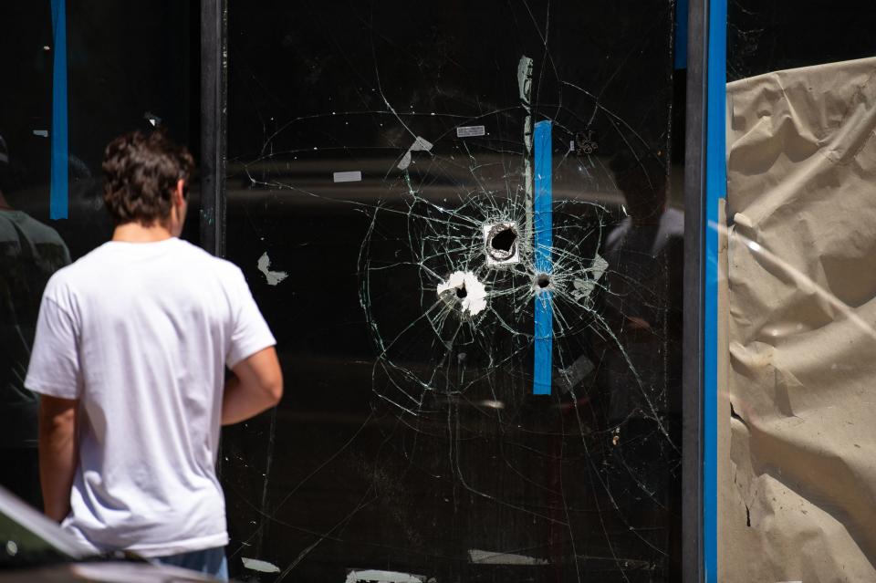 A man walks past bullet holes in the window of a storefront.