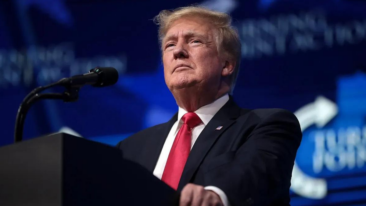 A white man wearing a black suit stands at a podium. 