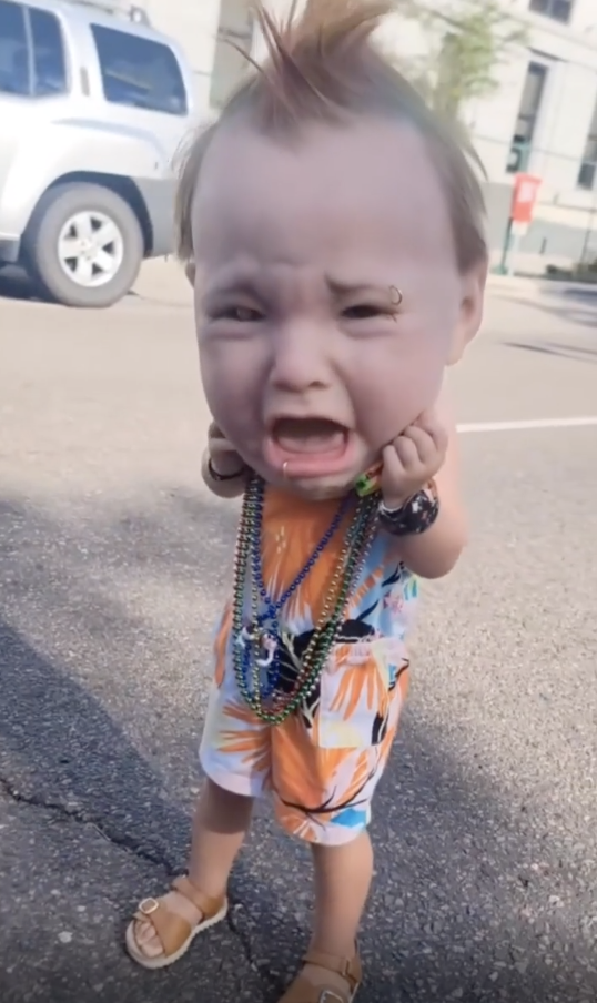 A child in a colorful outfit and sandals wearing several pearl necklaces stands outside with a comically exaggerated, desperate expression on his face