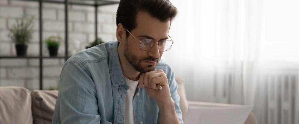 Thoughtful young caucasian man in glasses sit on sofa in living room read postal letter