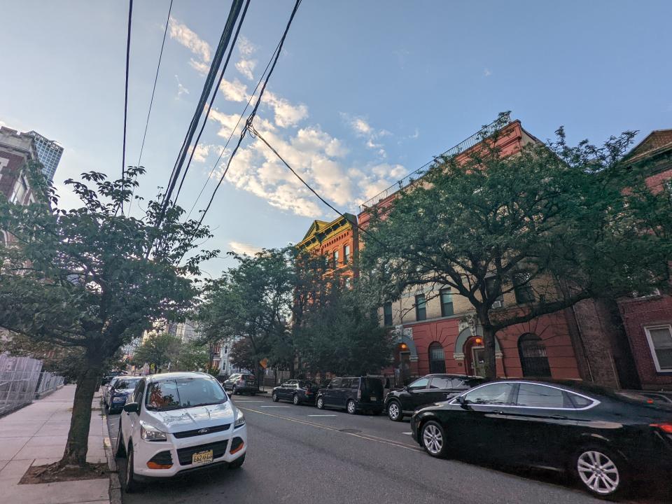 <p>A picture from the Pixel 6a's ultrawide camera, featuring a red building behind some trees in front of a road lined with cars on both sides. In the background is a blue sky and some clouds.</p>
