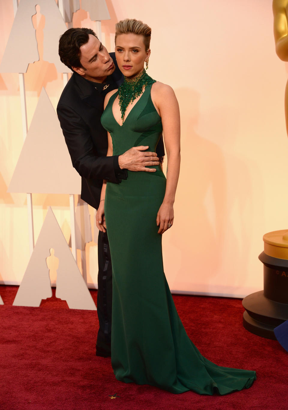 Scarlett Johansson stands on the Oscars red carpet in a green gown, John Travolta stands behind her with his hand on her stomach, he's leaning in to kiss her cheek