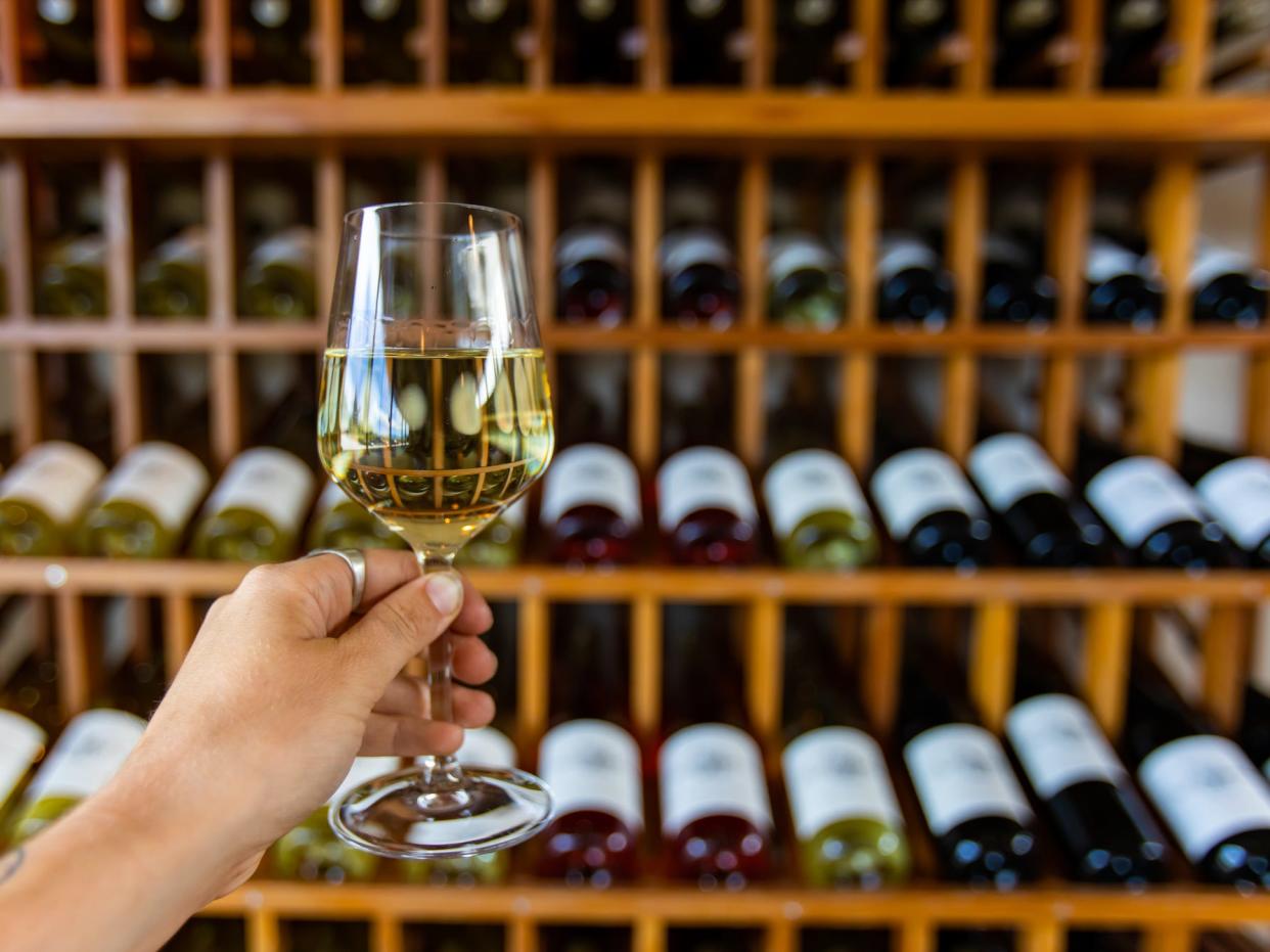 hand holding a glass of white wine in front of racks of wine bottles