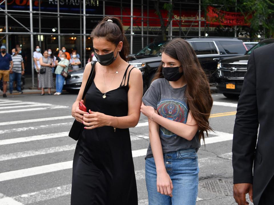 Katie Holmes, in a black sleeveless dress and black mask, walks across the street with daughter Suri Cruise, in a gray t-shirt, jeans, and black mask.