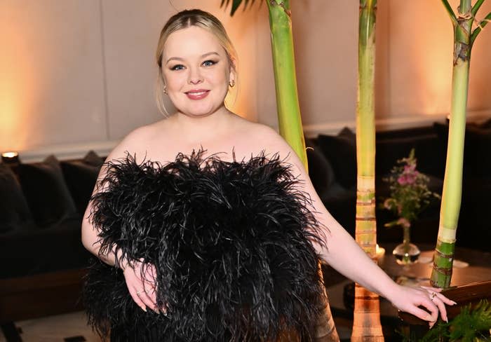 Nicole Byer at an event, in a black feathered dress, smiling, standing next to bamboo decor