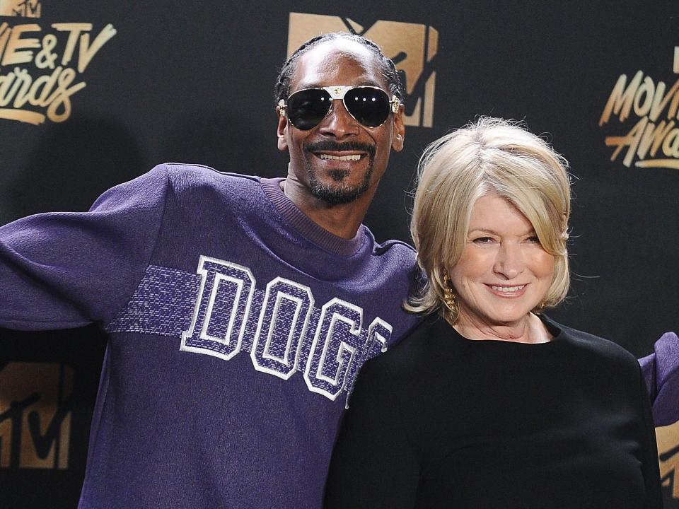 Snoop Dogg smiling with his arms wide open behind Martha Stewart smiling.
