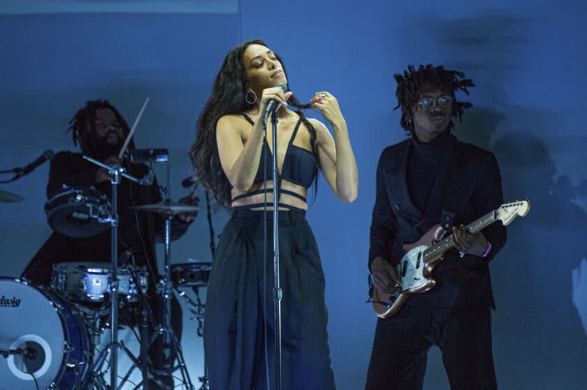 A woman with long black hair standing in front of a microphone in front of a band on a stage