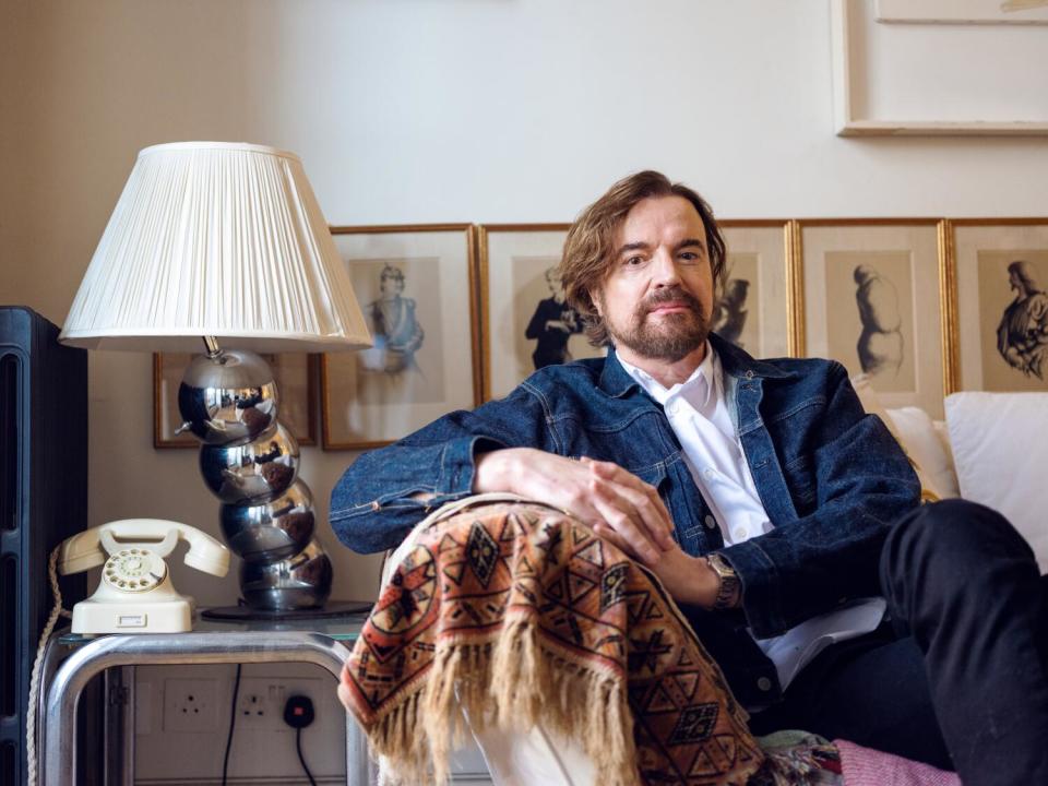 A bearded man sits on a sofa leaning against the arm for a portrait.