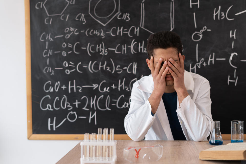 A teacher sits in a white coat against the background of blackboard with formulas, covering his face with his hands