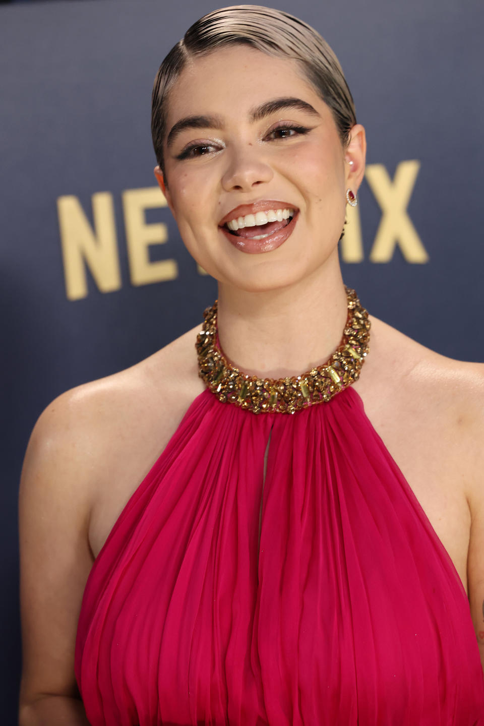 Auli'i Cravalho in pleated gown with gold neckpiece smiling against event backdrop
