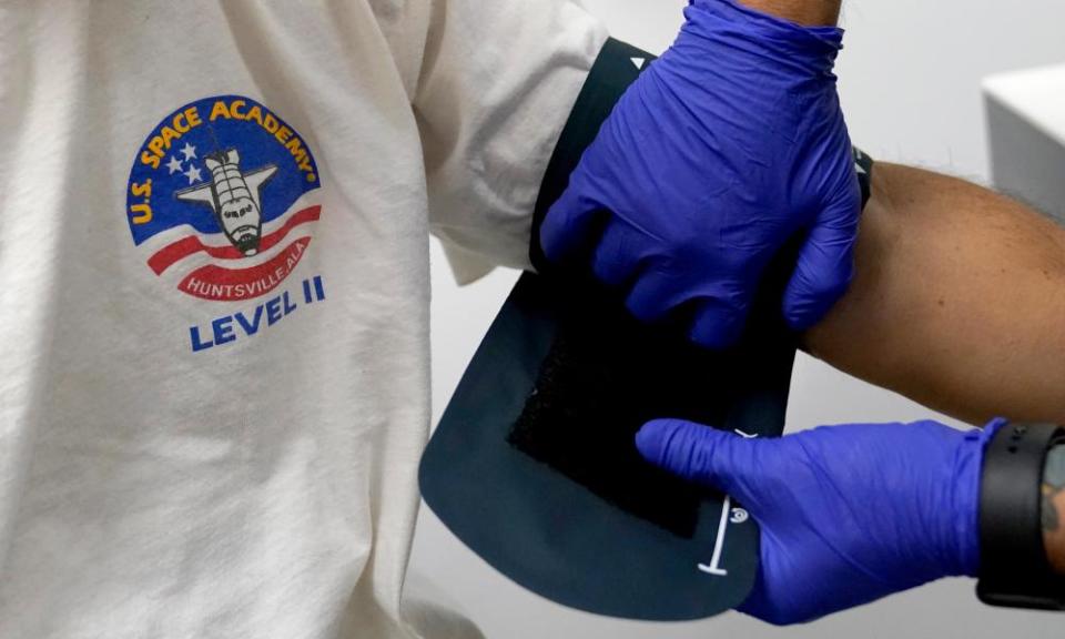 A close-up of a white T-shirt with a US Space Academy logo on it, and hands in purple glove arranging a black blood-pressure cuff on a man’s arm.