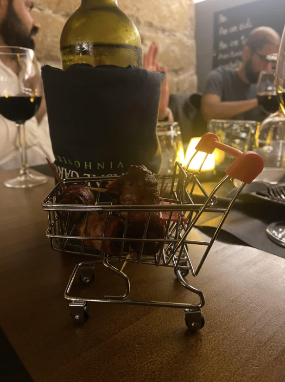 Miniature shopping cart filled with appetizers on a table at a restaurant, with blurred individuals dining in the background