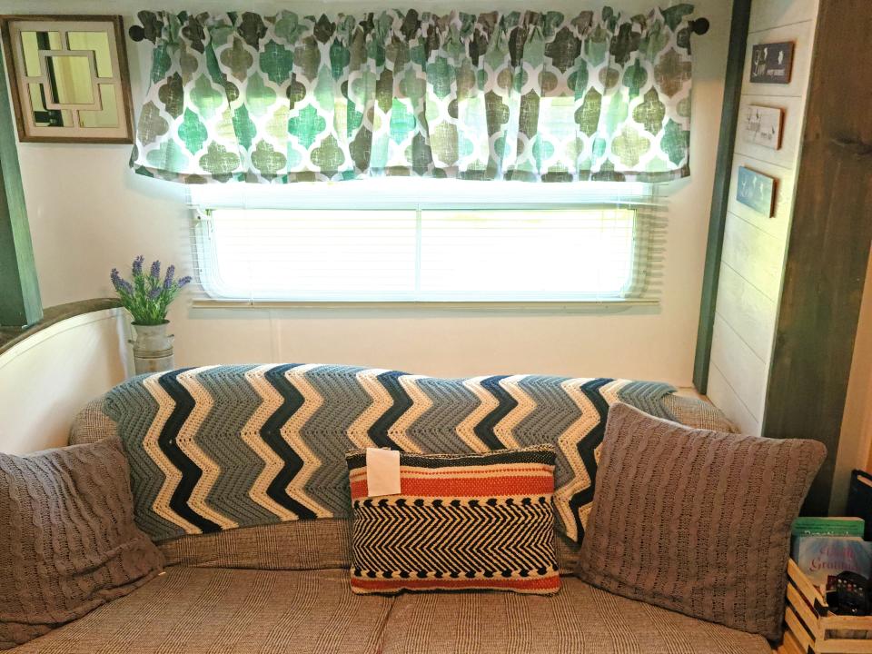 A grey couch with two grey pillows, one patterned pillow, and a blue and white zig-zag-patterned blanket. There are decorative signs on the wall and a window with a short curtain.