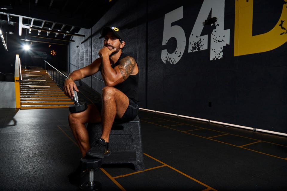 54D founder Rodrigo Garduño sits on a plyo box with two dumbbells in an empty gym with large letters reading "54D" on the wall behind him.