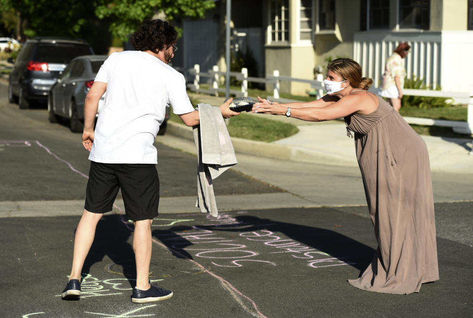 In this May 9, 2020 photo, musician Adam Chester, left, receives carrot cake cupcakes from neighbor Caryn Adams over a social-distancing chalk line before Chester's weekly neighborhood concert in the Sherman Oaks section of Los Angeles. Normally, Chester is a surrogate Elton John, who sings and plays the rock superstar's parts at rehearsals. With that work on hold, Chester has been giving concerts to his neighbors from a safe social distance in front of his house. (AP Photo/Chris Pizzello)