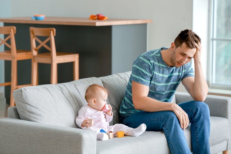 A frustrated and sad father sits on the couch next to his baby.