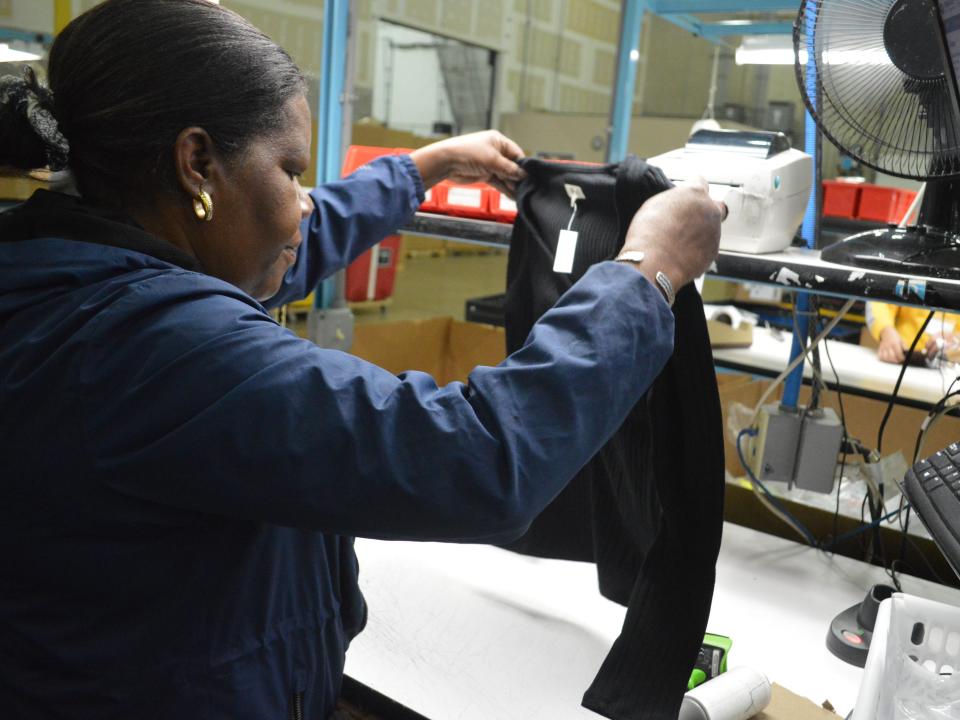 A black woman in a blue hoodie inspects a black sweater inside a returns processing warehouse.