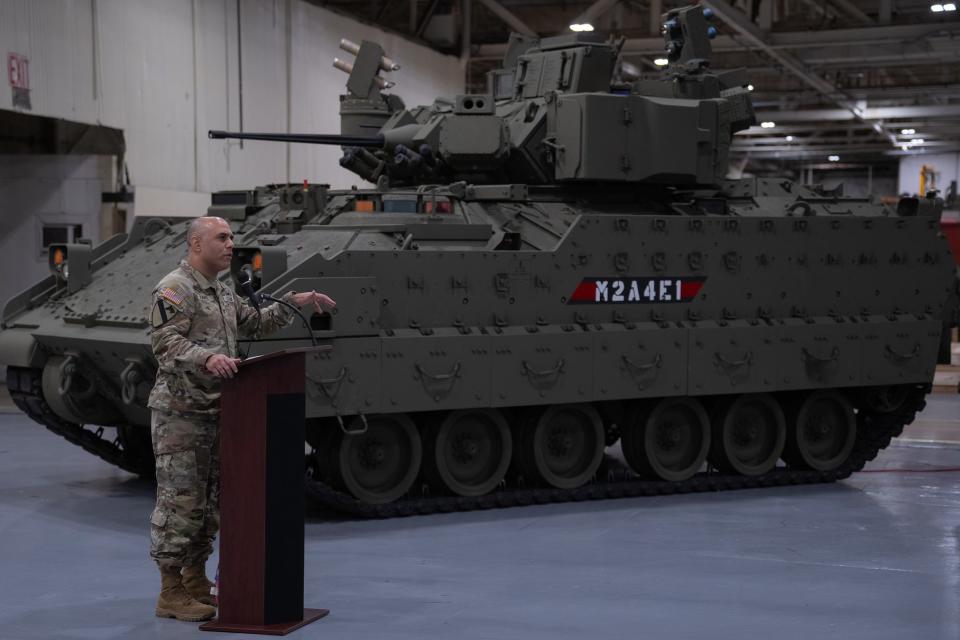 A member of the US Army speaking from a podium next to an infantry fighting vehicle.