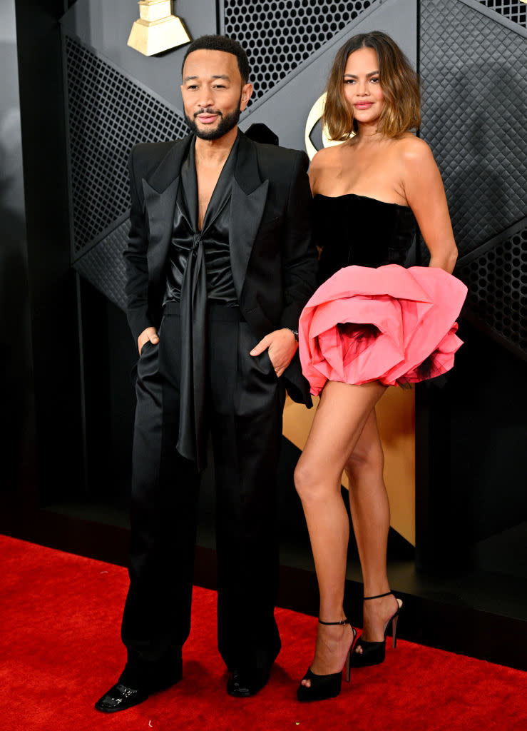the couple on red carpet, John in suit and Chrissy in short dress with large ruffle