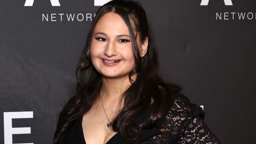 gypsy rose blanchard smiles at the camera while standing in front of a black background with white writing, she wears a black vneck dress and a necklace