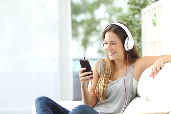 A young woman listens to music on headphones through her smartphone on a couch.